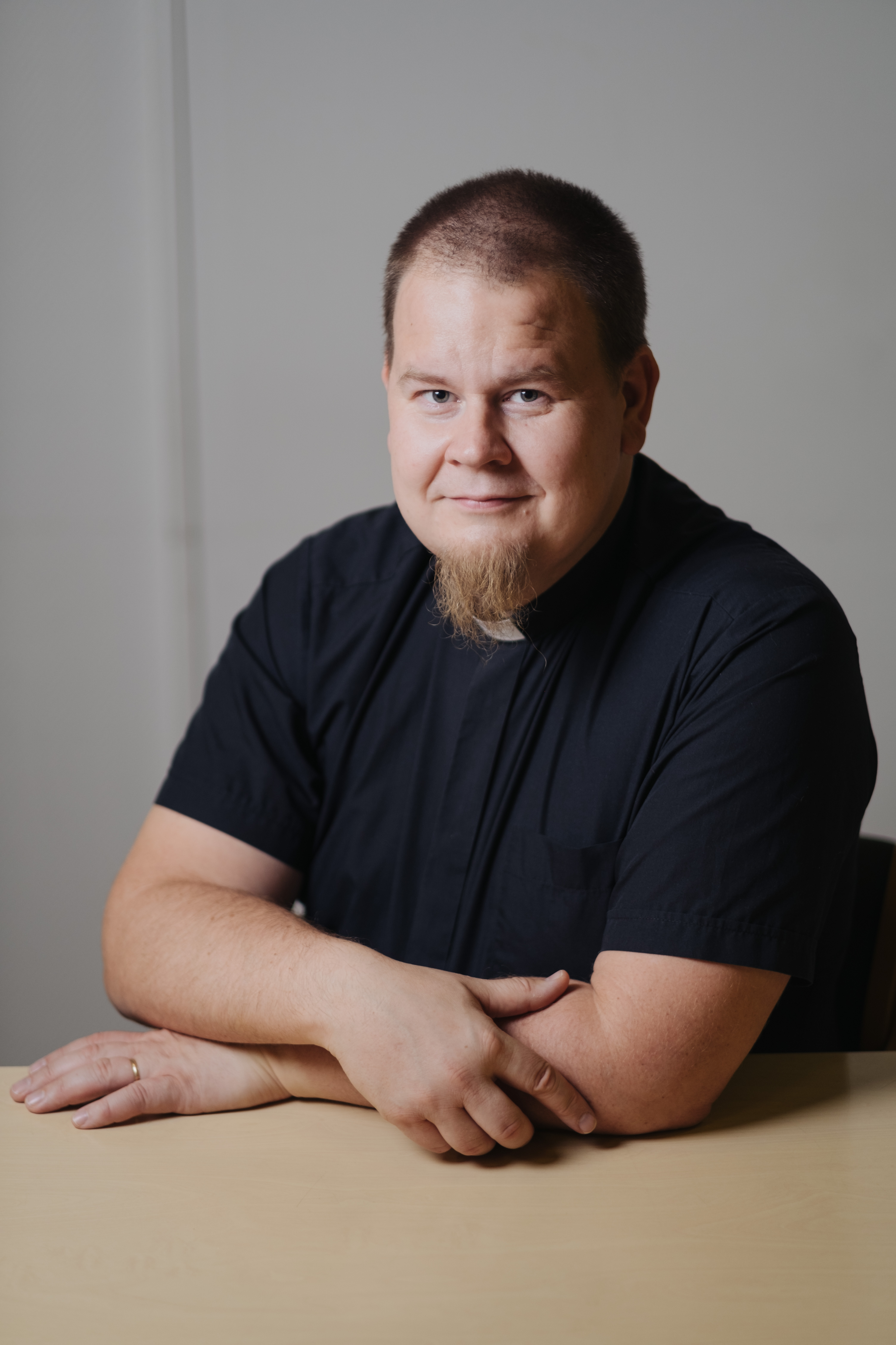Hiltunen on pukeutunut mustaan, lyhythihaiseen papinpaitaan ja nojaa pöytää vasten.