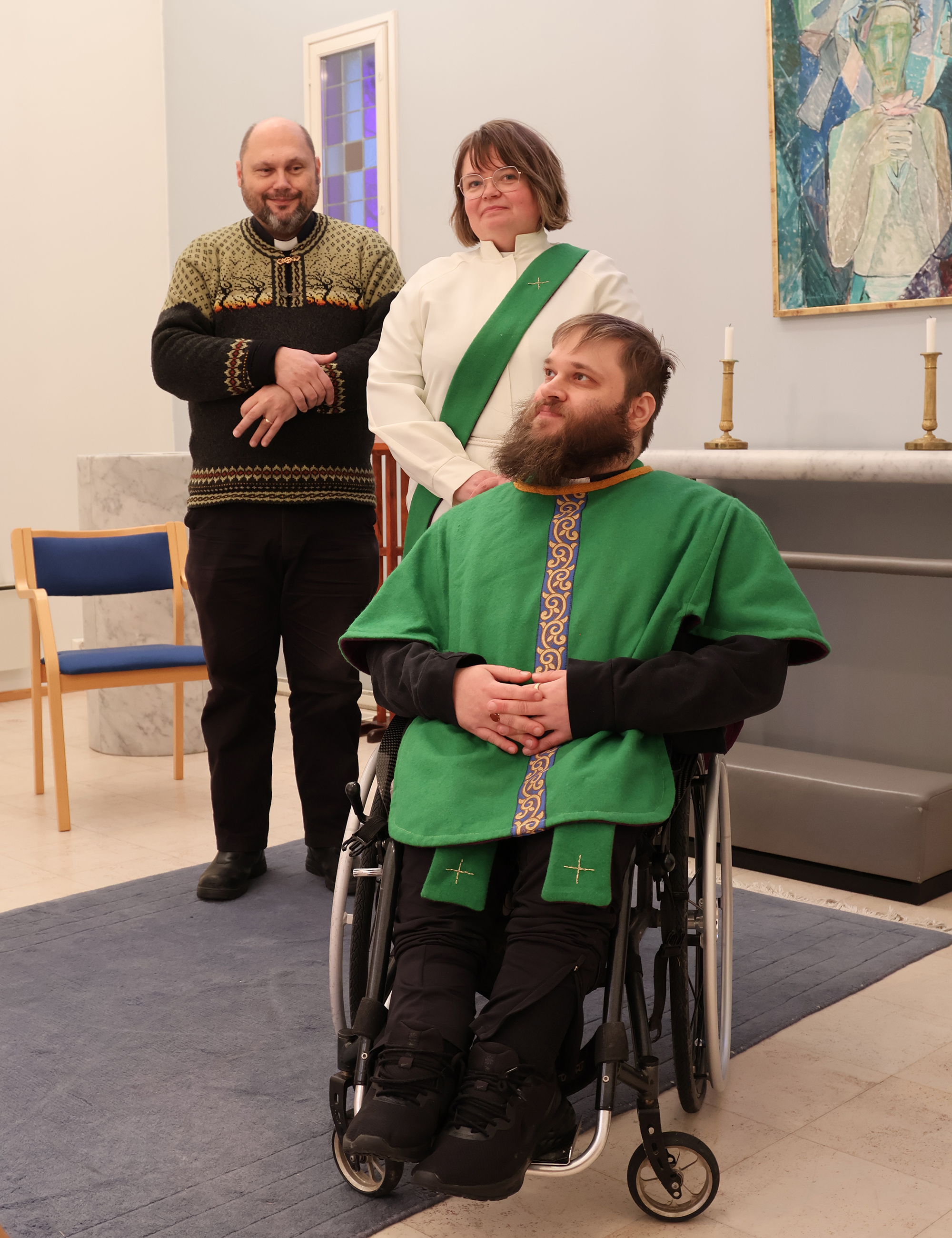 Kaksi ihmistä seisoo ja yksi istuu pyörätuolissa alttarin edessä.