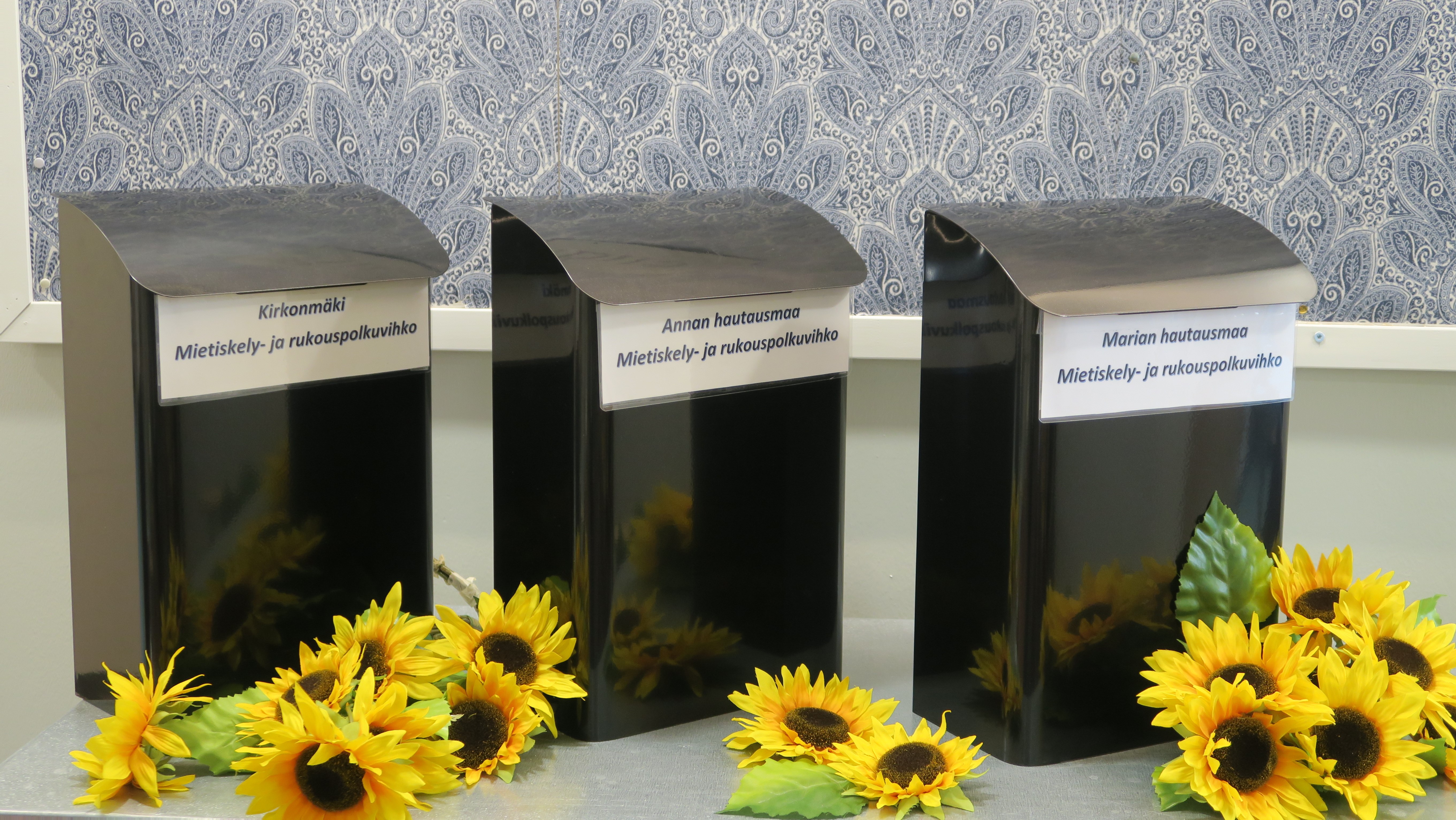 Kolme metallista postilaatikkoa rivissä aurinkokukkia koristeena.